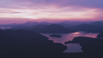 Violett-pinker Sonnenuntergang über einer bergigen Landschaft mit Wasserflächen und silhouettierten Hügeln.